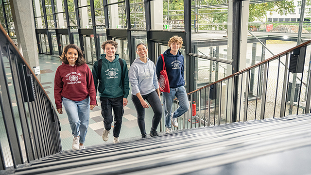 Vier Studierende gehen nebeneinander eine Treppe hoch, im Eingangsbereich des Audimax. Das Foto ist von oben an der Treppe aufgenommen.